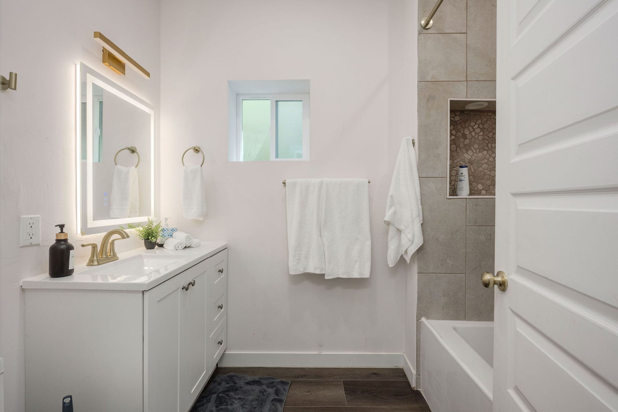Bath with white vanity, lighted mirror, brass fixtures, and tiled tub-shower with pebble niche