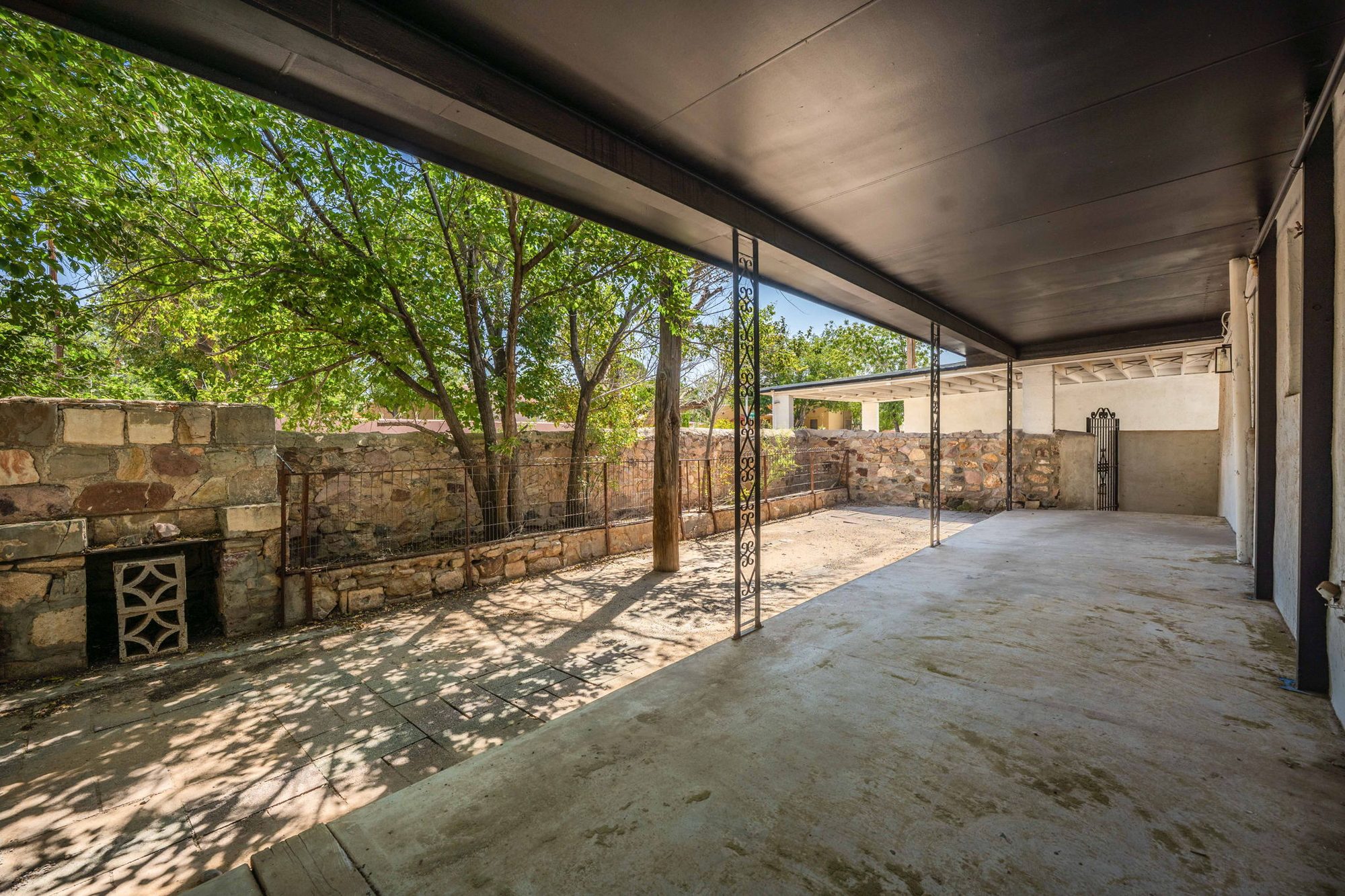 Long covered patio with ironwork posts beside a stone-walled yard under shade trees